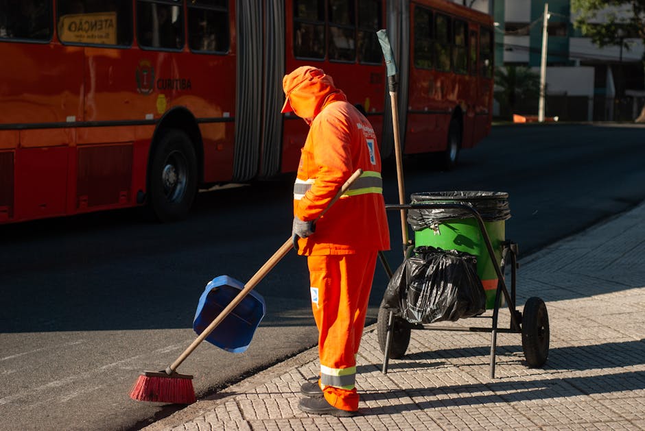 A professional cleaner wearing a black beanie, gray hoodie, and blue jeans is engaged in surface cleaning outside a modern building, using a squeegee and spray bottle on large glass panels. The glass 